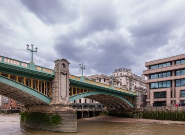 London, England, UK - July 6, 2022: From Thames River. Southwark bridge landing on north shore under heavy dark cloudscape. Buildings on both sides.