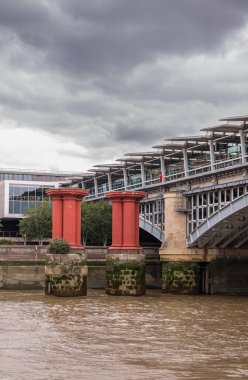 London, England, UK - July 6, 2022: From Thames River. Oxblood red pillars protecting Blackfriars railway bridge NW shore under thick gray cloudscape.