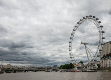 London, England, UK - July 6, 2022: From Thames River, Westminster pier. Wide view, The London Eye against gray cloudscape. Hungerford and Golden Jubilee bridges on horizon.