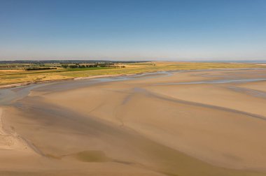 Mont St. Michel, Normandiya, Fransa - 8 Temmuz 2022: Mavi gökyüzünün altındaki kayanın batısındaki ebb ıslak kumlu plaj manzarası. Yeşil-sarı bataklık arazisi ve ufukta yeşil ağaç çizgisi.