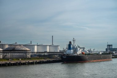 Rotterdam, Hollanda - 11 Temmuz 2022: Kriti Kaya petrol tankeri akşam gökyüzünün altındaki petrol ve gaz depolama tanklarının önünde. Rafineri bacası arkada.