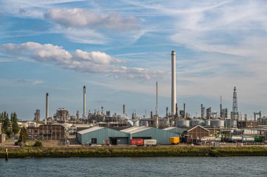 Rotterdam, Hollanda - 11 Temmuz 2022: Oude Maas nehri kıyısındaki depoların önünde kamyonlarla mavi akşam bulutları altında kabuk Pernis rafinerisi