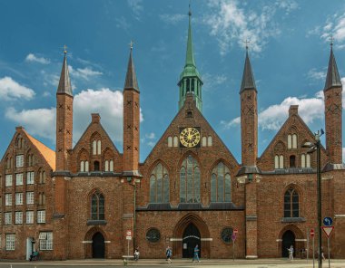 Almanya, Lubeck - 13 Temmuz 2022: Kahverengi tuğlalı Heiligen Geist Hastanesi ön cephesi önü mavi bulutların altında sokak manzaralı. Kuleler ve altın saat