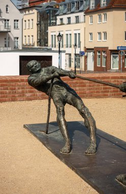 Almanya, Wismar - 13 Temmuz 2022: St. Marienkirche. Tauziehen 'deki bir adam ya da ön taraftaki halat çekme heykeli. Arkadaki meydanın ön cephesinde çok renkli evler var. Sarı çakıl tabanı. 