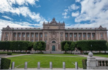İsveç, Stockholm - 16 Temmuz 2022: Doğu Cephesi, Bej Taşı Forsvarsutskottet veya Riksdagshuset veya Parlamento yeşil parkın arkasındaki mavi bulutların altında. Gable 'daki heykeller