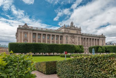 İsveç, Stockholm - 16 Temmuz 2022: Doğu Cephesi, Bej Taşı Forsvarsutskottet veya Riksdagshuset veya Parlamento yeşil parkın arkasındaki mavi bulutların altında. Gable 'daki heykeller