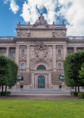 İsveç, Stockholm - 16 Temmuz 2022: Doğu Cephesi, Bej Taşı Forsvarsutskottet veya Riksdagshuset veya Parlamento yeşil parkın arkasındaki mavi bulutların altında. Gable 'daki heykeller