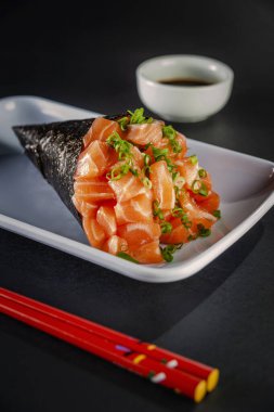 Salmon temaki on white plate in black background with soy sauce and chopsticks. Closeup.