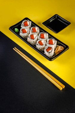 Uramaki salmon with soy sause and chopsticks in a black and yellow background.