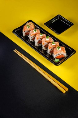 Uramaki blowtorcherd salmon with soy sause and chopsticks in a black and yellow background.