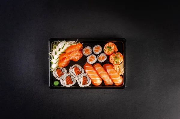 Tray sushi salmon varieties for delivery: sashimi, onigiri, sushi Jhow, sakemaki and uramaki philadelphia on a black background. Top view.