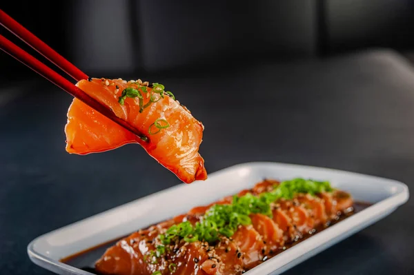 Salmon sashimi on chopsticks with sesame, shoyu and miso sauce on a black table. Closeup.