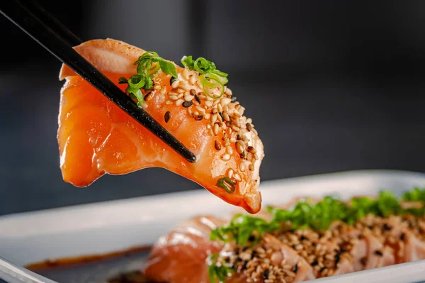 Blowtorcherd salmon sashimi on chopsticks with sesame, shoyu and passion fruit sauce on a black table.