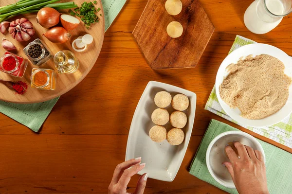 Kadın elleri Brezilya kroketini (coxinha de frango) ahşap mutfak masasında ekmek kırıntılarıyla hazırlıyor.