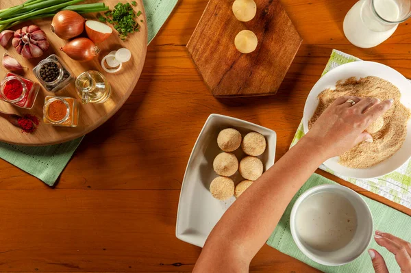 Kadın elleri Brezilya kroketini (coxinha de frango) ahşap mutfak masasında ekmek kırıntılarıyla hazırlıyor.