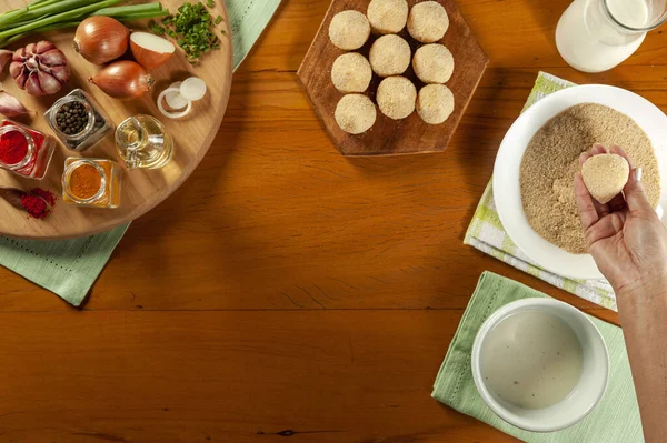 Kadın elleri Brezilya kroketini (coxinha de frango) ahşap mutfak masasında ekmek kırıntılarıyla hazırlıyor.