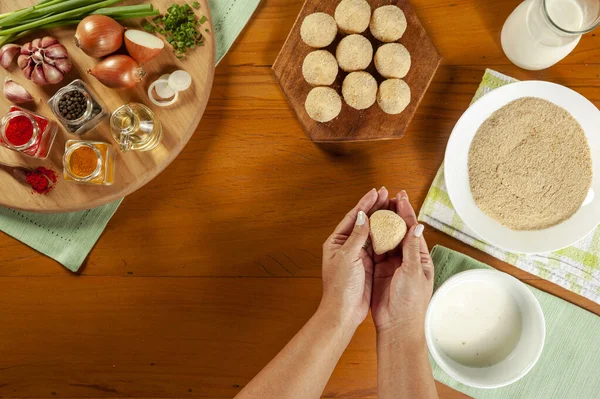 Kadın elleri Brezilya kroketini (coxinha de frango) ahşap mutfak masasında ekmek kırıntılarıyla hazırlıyor.