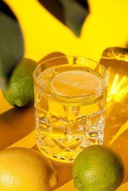Sarı ve yeşil limonlu limon dilimli, sarı arka planda gölgeli bir bardak su. Yaz içeceğini tazeliyorum.