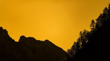 Ağaçların siluetleri dağların arka planına ve turuncu gökyüzüne karşı. Valbona Valley Ulusal Parkı. Prokletije Dağları. Arnavut Alpleri.