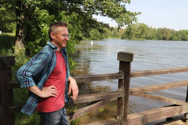 Sırt çantası ve büyük tahta bir köprüyle nehir kıyısında duran genç adam.