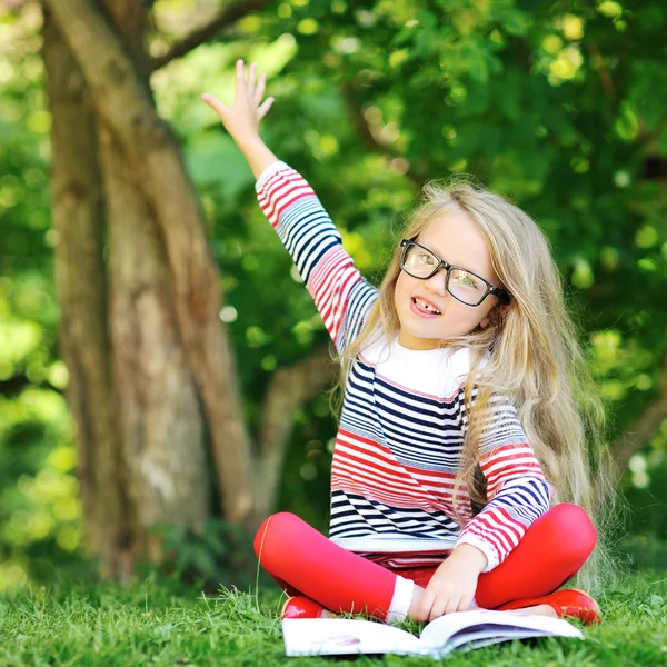 Little girl with a book in a park wearing glasses pointing at co — Stock Photo, Image