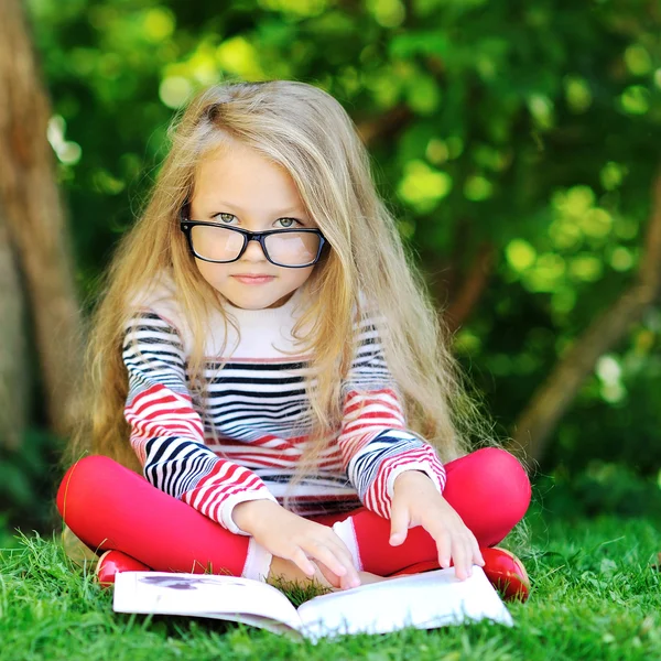 Sweet little girl wearing glasses and reading book in a park — Stock Photo, Image