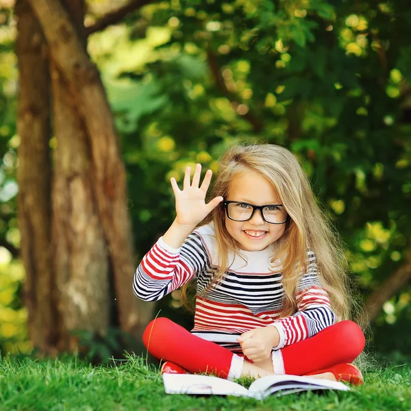 Cute little girl sits in a park. Reading book and hand gesture w — Stock Photo, Image