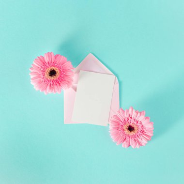 İçinde pembe zarf, boş kart ve mavi pastel arka planda bahar çiçeği aranjmanı olan minimal kompozisyon. Düz yatış, üst görünüm.