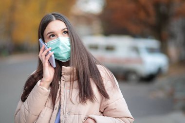 Tıbbi maskeli bir kadın cep telefonuyla ambulansın arka planında konuşuyor. COVID-19 koronavirüs salgını.
