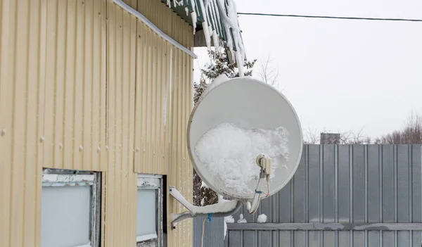 降雪後の雪の中の家の壁に固定された衛星料理 ストック写真