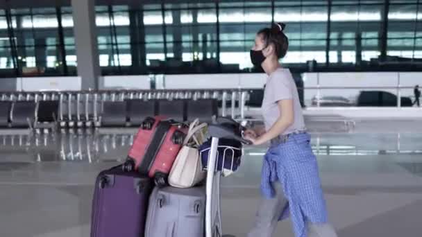 顔マスクをした若い女性が空港ターミナルで多くの袋で荷物トロリーを押してチェックインする前に歩く。流行と旅のコンセプト — ストック動画