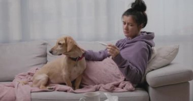 Genç hasta bir kadın termometreyle kanepede küçük köpeğiyle yatıyor, mevsimsel grip ya da soğuk algınlığından muzdarip. Hasta kız evde grip olunca kendini kötü hisseder.