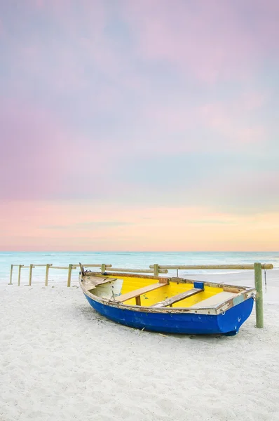 Старая желто-голубая деревянная лодка на белом пляже на закате — стоковое фото