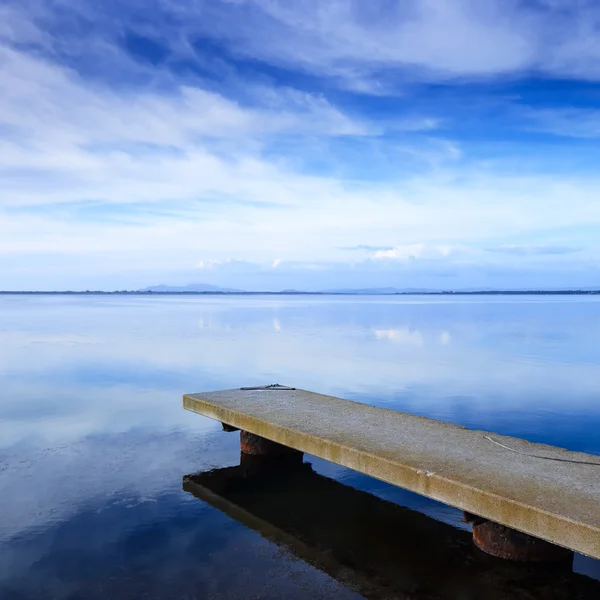 Konkreta brygga eller brygga och en blå sjön och sky reflektion över vatten. — Stockfoto