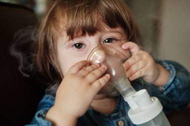 Maskeli küçük bir kız, evde nebulizörle solunum yolu tedavisi görüyor. Bebek ağzında nebulizör, astım spreyi ve bronşit tedavisi ile oturuyor. Nebulizör, astım spreyi kullanan tatlı bir kız..