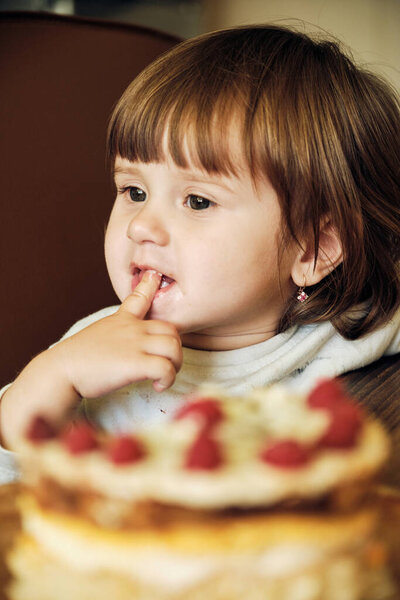 Cute Happy Baby Girl Eating Cake With Raspberries. Adorable little girl Tasting Cream On Cake. Closeup