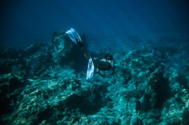 Sualtı ateş eden kız, temiz suda siyah bir mayo giyerek suya dalıyor.