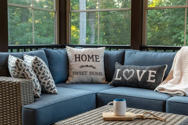 A cozy corner of an outdoor living space with modern porch windows, patio furniture, cushions and pillows. Woods in the background.