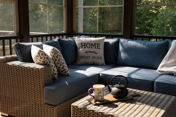 A cozy corner of an outdoor living space with modern porch windows, patio furniture, cushions and pillows. Coffee or tea pot and cups on coffee table, woods in the background.