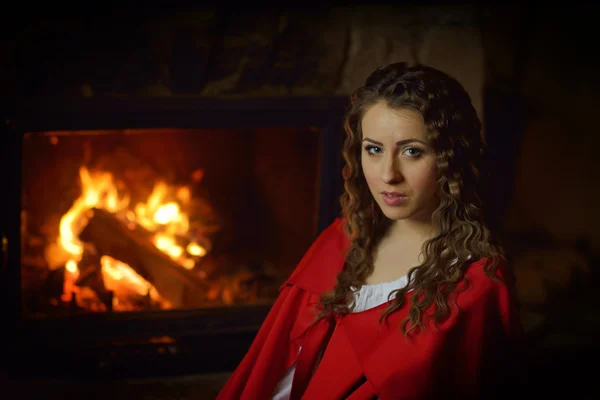 Piękna kobieta z czerwony płaszcz, siedząc przy kominku — Zdjęcie stockowe