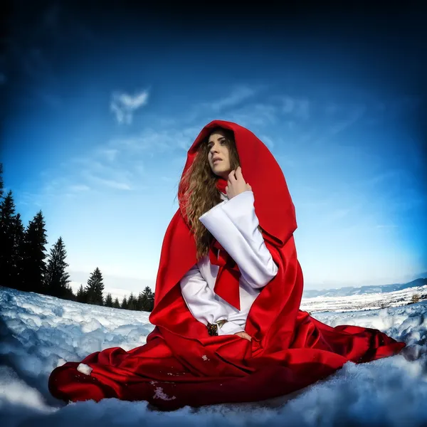 Mulher bonita com manto vermelho sentado na neve no inverno — Fotografia de Stock