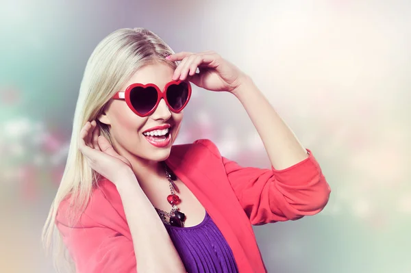 San Valentino ritratto di una giovane ragazza bionda attraente con occhiali a forma di cuore ben vestito indossando abito viola sullo sfondo rosa Fotografia Stock