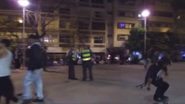 İnsanlar geceleri meydanda, polis memurlarının yakınında kayıyor ve yürüyor.