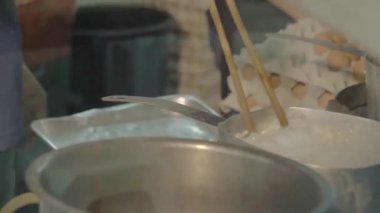asian man cooking noodles in a big silver pot with huge chopsticks