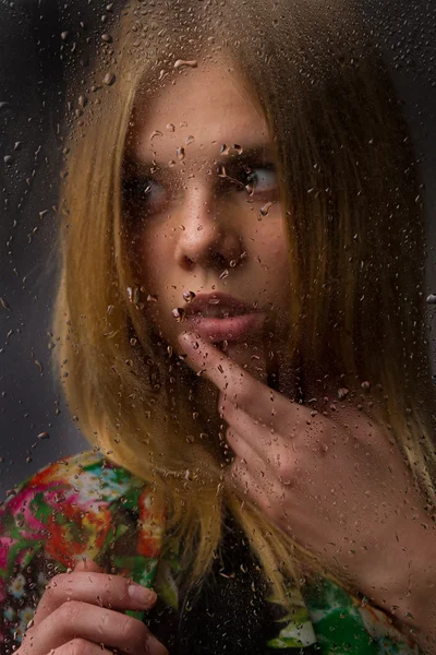 Retrato de uma linda garota atrás de vidro molhado em um fundo preto . — Fotografia de Stock