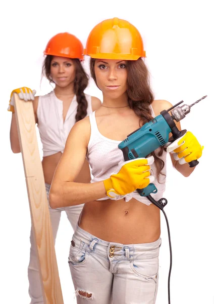Gêmeos meninas em capacetes laranja com uma broca elétrica e serra elétrica em um fundo branco . — Fotografia de Stock