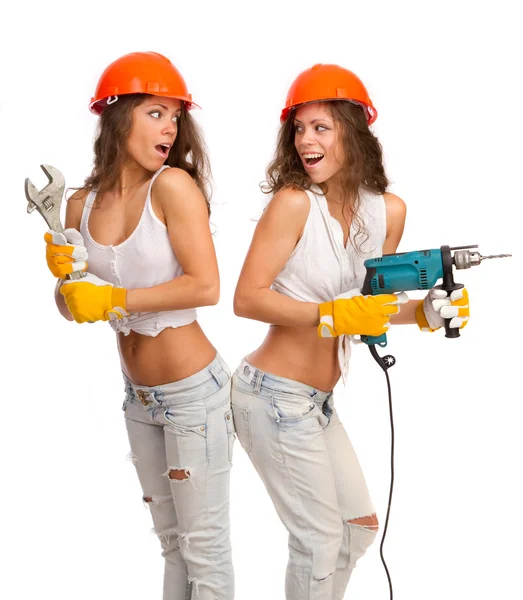 Gêmeos meninas em capacetes laranja com uma broca elétrica e serra elétrica em um fundo branco . — Fotografia de Stock