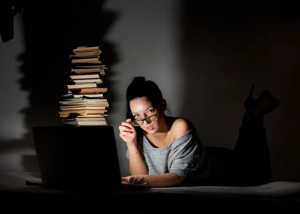 Bella studentessa sdraiata sul pavimento con laptop e libri su uno sfondo scuro — Foto Stock