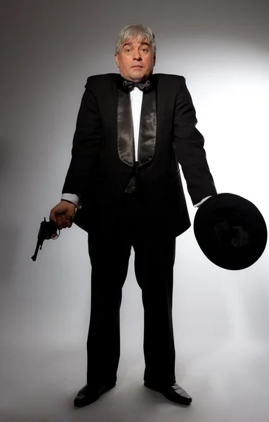 The elegant man in a tuxedo with a hat and a gun on a gray background — Stock Photo, Image