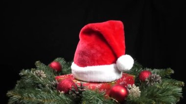 Noel Baba şapkası yeşil Noel ağacı dalları, koniler ve Noel toplarıyla çevrili.